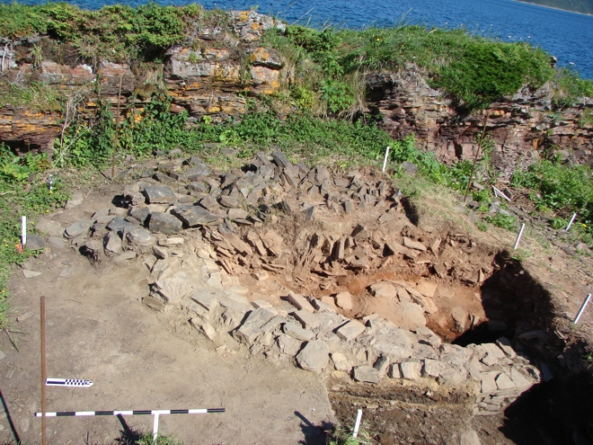The excavation of the bread oven at Champ Paya (EfAx-09, Feature 22), Cape Rouge Harbour. The outer wall of the East side can be seen at the bottom of the image. The orange/brown deposits mark the interior of the oven where baking took place. The oven had been deliberately collapsed when abandoned. Note how the oven is situated in the shelter of a natural bedrock outcrop.